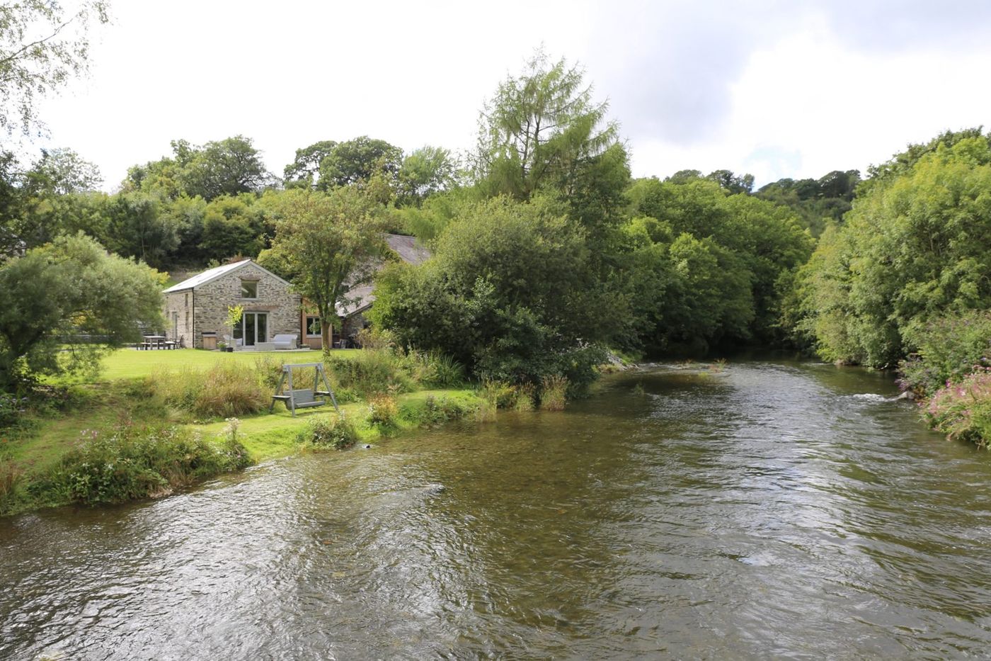 The Cottage seen from across the river, stone walls among trees with the lawn running down to the water