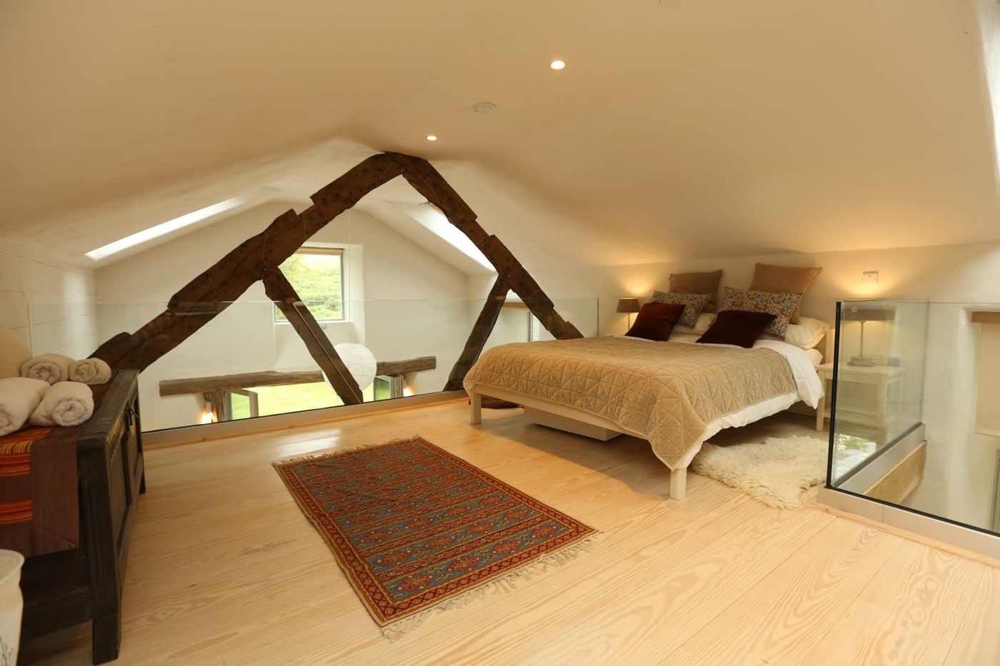 The mezzanine bedroom under the roof beams, with a glass balustrade and a rug on the pale wooden floor