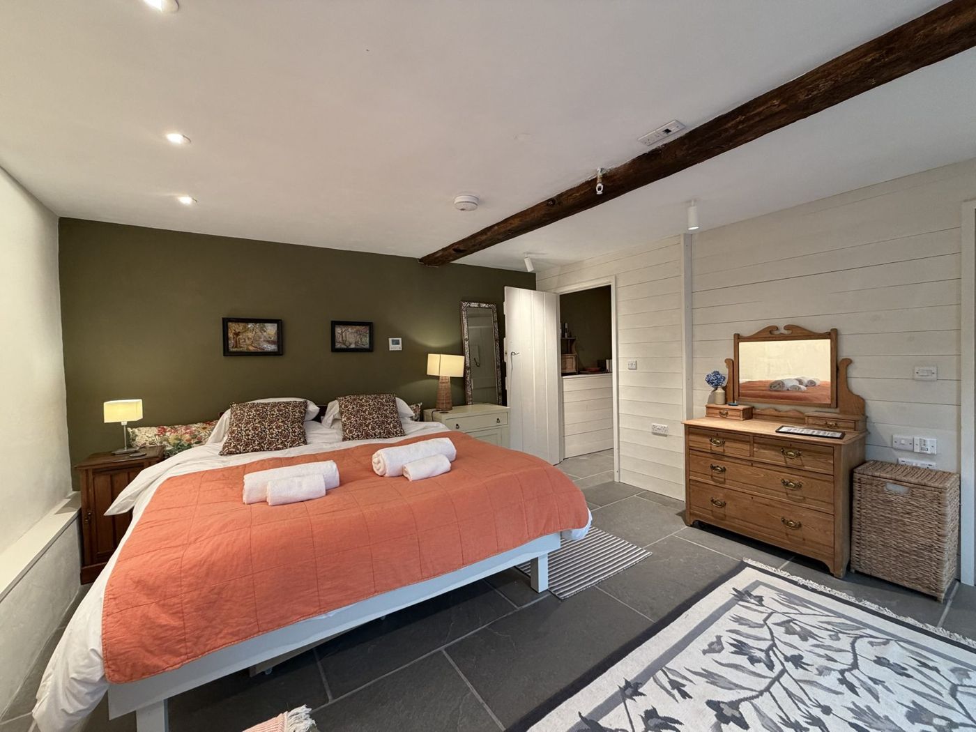 The bedroom: a double bed with an orange quilt, olive wall, oak beam and pine chest of drawers